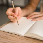 La photo d'une main tenant un crayon et se préparant à écrire sur un cahier, symbolisant le fait d'écrire le livre de sa vie